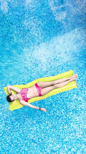 Beautiful young woman in hat in swimming pool aerial top view from above, young girl in bikini relaxes and swims on inflatable mattress and has fun in water on family vacation, tropical holiday resort - Photo, Image