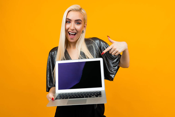 serious charming blond woman shows a blank laptop screen with a mockup in front of herself against a yellow wall with copy space - Foto, Bild