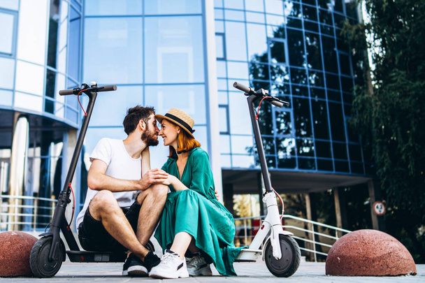 Young loving couple relaxing sitting near a modern glass building with their electric scooters. Walks in the big city. - Foto, Imagen