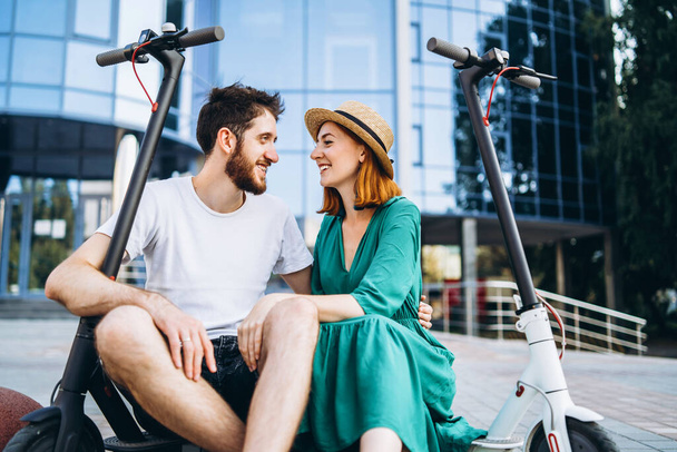 Couple of two attractive people are chilling near the glass building with their electro scooters. Man and woman enjoy vacation. - Foto, Imagen