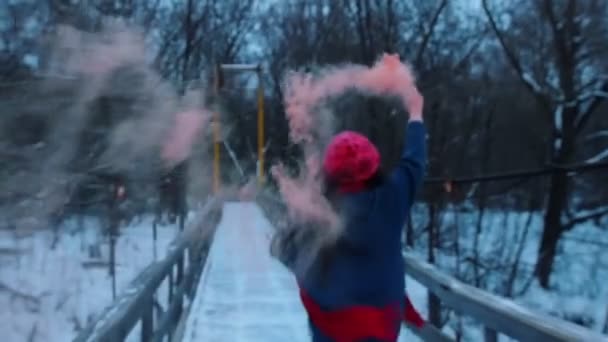 Een jonge vrouw op de besneeuwde brug met een roze rookbom. - Video