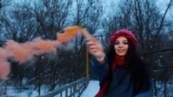 A young beautiful woman standing on the snowy bridge holding an orange smoke bomb - Πλάνα, βίντεο