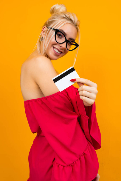 Europäisches Mädchen in einem roten eleganten Kleid mit Kreditkarte zum Einkaufen in der Hand mit einer Attrappe auf gelbem Studiohintergrund - Foto, Bild