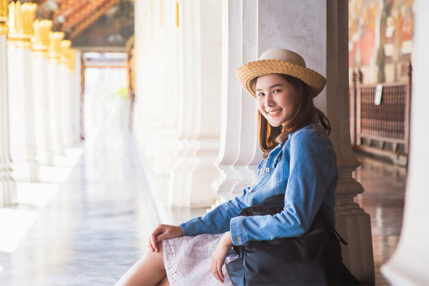 Medium close up portrait smiling young Asian woman in jeans jacket and straw hat and backpack at side, sitting and resting on parvilion floor at Wat Phra Kaew Temple. - Fotografie, Obrázek