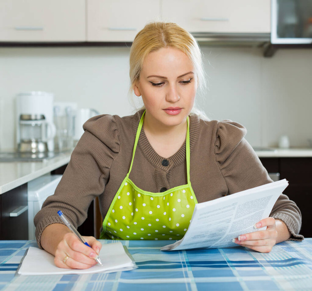 Serious woman filling in financial documents at table in home kitchen  - Photo, Image
