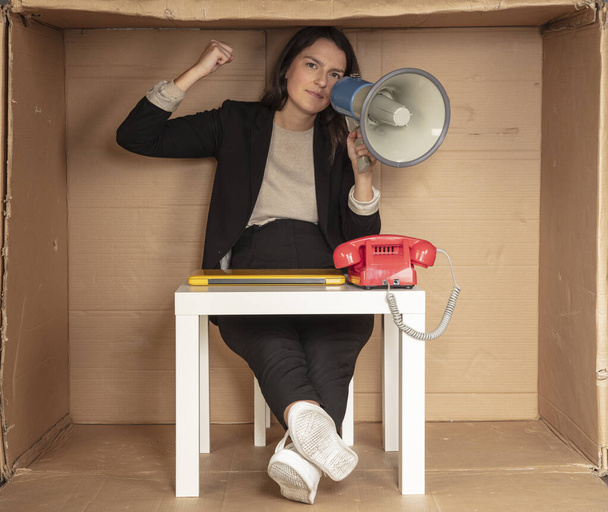 business woman with a megaphone in hand sits in a cramped cardboard office - Fotografie, Obrázek