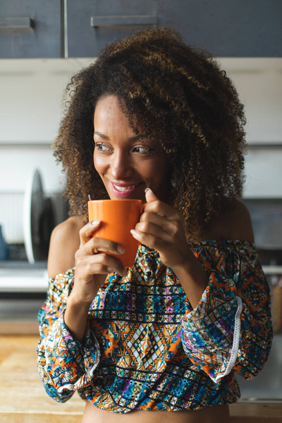 Mujer negra joven casual tomando una relajante taza de café en la cocina en casa. Modelo de peinado afro
. - Foto, Imagen