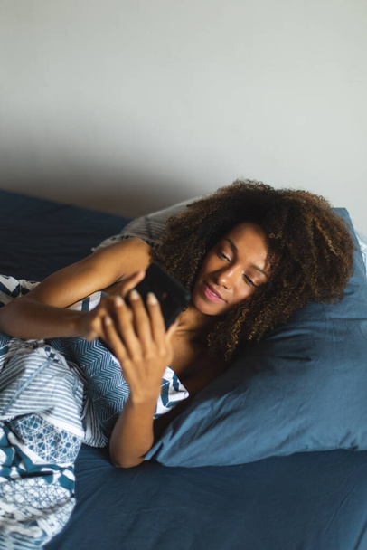Afro hairstyle young woman in bed waking up and looking her smartphone for checking messages and social networks. - Foto, Imagem