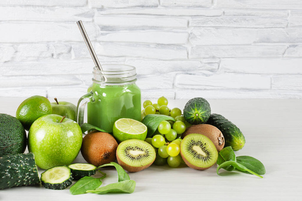 Various green fruits and vegetables with mason jar of smoothie on white wooden table over white brick wall. Detox diet. Healthy eating concept. - Photo, Image