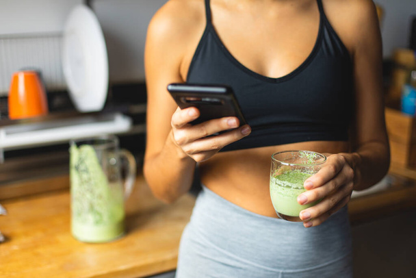 Fitness diet concept. Sporty woman holding a green detox smoothie and using nutrition app on smartphone in the kitchen. Fit breakfast. - Fotó, kép