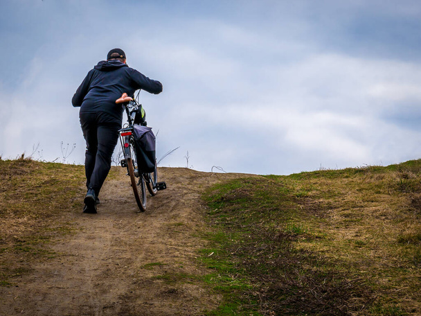 男が自転車で丘を歩いている。曇りの日に男と彼の自転車. - 写真・画像
