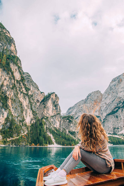 Young tourist woman, long wavy hair turned away back sits in wooden boat trip. Casual clothes warm brown sweater enjoy silence dream relaxation luxury nature Italian Alps backdrop fabulous lake Braies - Foto, imagen