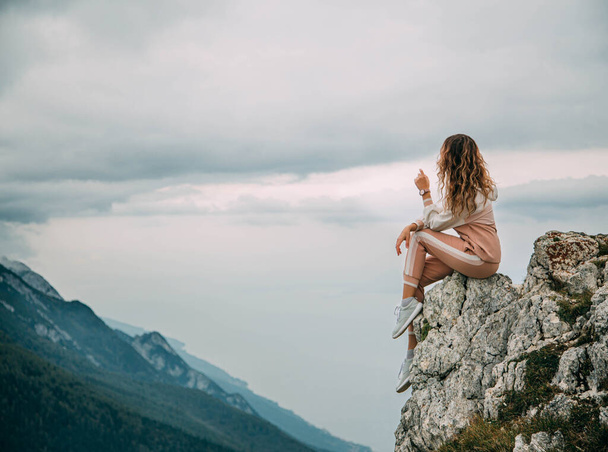 Red hair girl turned away in pink suit sits on edge mountain rock on backdrop sky clouds mountains foggy haze. Woman enjoy beauty fairytale nature silence relaxation. Top Peak Monte Baldo Alps Italy - Zdjęcie, obraz