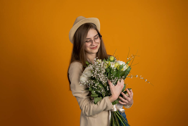 Charming young woman in hat and glasses holding flowers on yellow background - Foto, Imagem