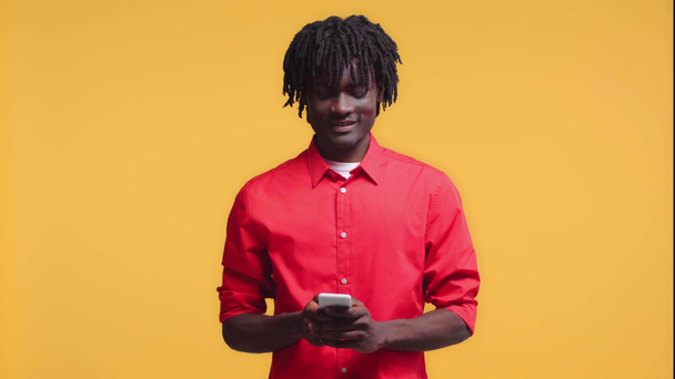 Afričan Američan chatování na smartphone a usmívá izolované na žluté - Záběry, video