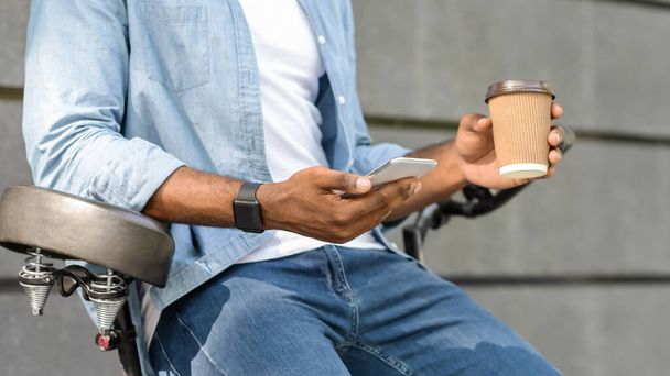 Musta kaveri istuu pyörän päällä, käyttää älypuhelinta ja juo kahvia ulkona
 - Valokuva, kuva