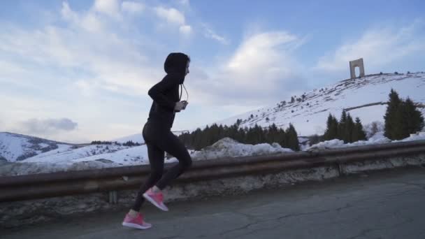 Siyah kapüşonlu ve pembe ayakkabılı bir kadın, karlı dağlarda koşuyor. - Video, Çekim