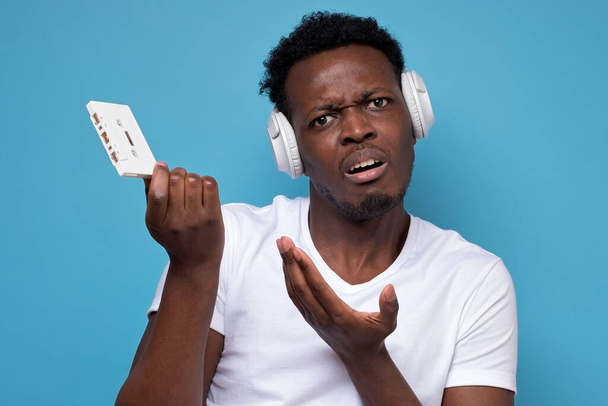 Beyaz kulaklıklı, elinde retro kaset olan genç, kafası karışmış Afrikalı bir adam onu mavi arka planda nasıl kullanacağını soruyor. Eski teknoloji konsepti. Stüdyo çekimi - Fotoğraf, Görsel