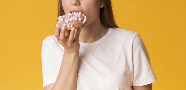 Hongerig onherkenbaar meisje eet donut, hongerig naar snoep op gele achtergrond - Foto, afbeelding