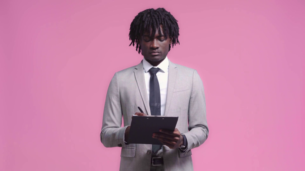 Empresario afroamericano escribiendo en portapapeles aislado en rosa - Metraje, vídeo