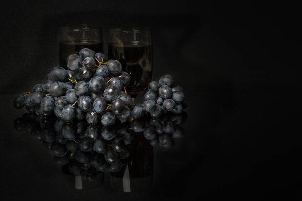 bunch of dark black grapes and two glasses of red wine on a dark mirror table with reflection. - Foto, Imagem