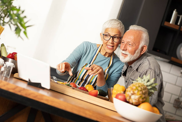 Modern technologies and seniors. Senior couple  reading recipe on tablet at kitchen, preparing healthy lunch - Foto, immagini