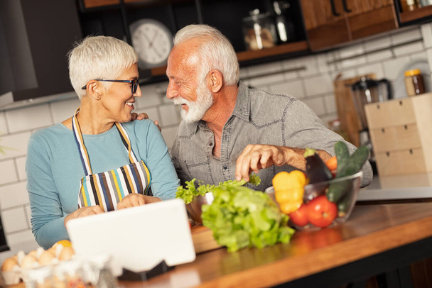 Happy senior couple cooking healthy meal together in home kitchen digital tablet helps them to prepare food - Fotoğraf, Görsel