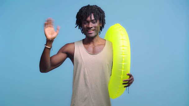 Hombre afroamericano pasando con anillo de natación y la mano ondulante aislado en azul
 - Metraje, vídeo