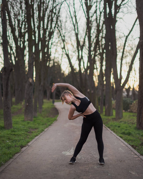 młoda blondynka dziewczyna z niebieskimi oczami i czarny strój robi na świeżym powietrzu ćwiczenia w parku w ciepłe zimowe popołudnie - Zdjęcie, obraz