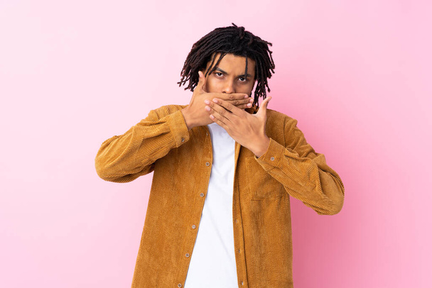 Jovem afro-americano com jaqueta de veludo sobre fundo rosa isolado cobrindo boca com as mãos
 - Foto, Imagem