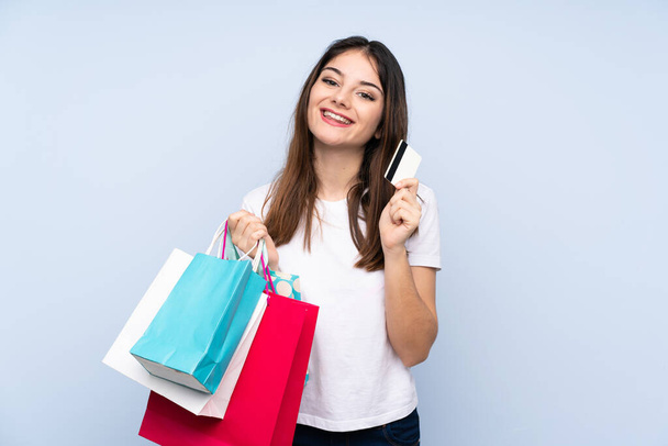 Mladá bruneta žena přes izolované modré pozadí držení nákupních tašek a kreditní karty - Fotografie, Obrázek