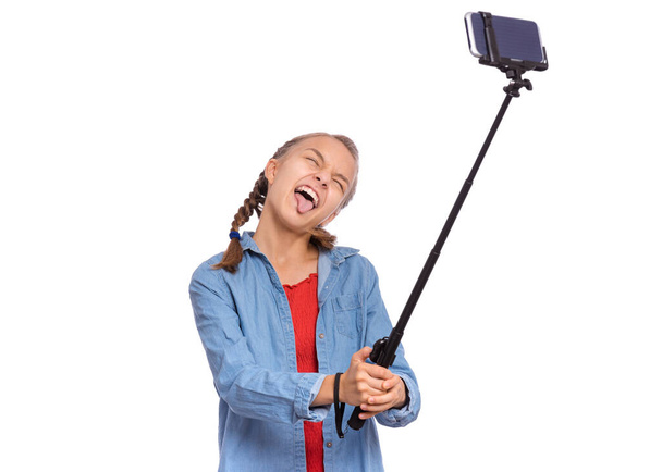 Portrait of happy teen girl making selfie with stick. Teenager posing to smartphone, making grimace - funny face, isolated on white background. Child taking selfie on mobile phone for social network. - Φωτογραφία, εικόνα