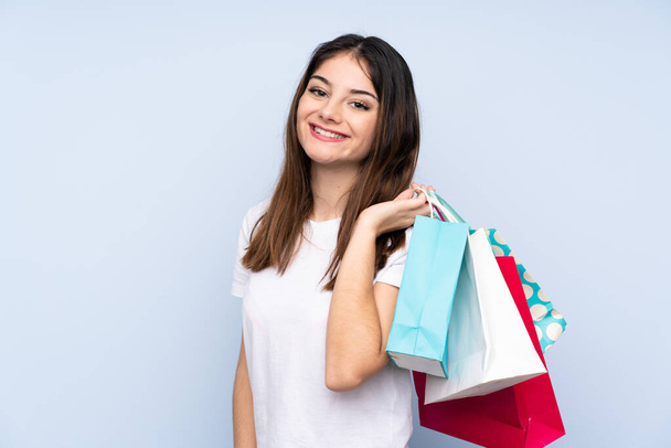Young brunette woman over isolated blue background holding shopping bags and smiling - Fotografie, Obrázek