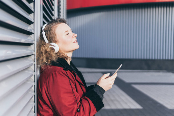 Young blonde woman listening to music in wireless headphones in the urban environment, holding mobile phone in her hands - Photo, Image