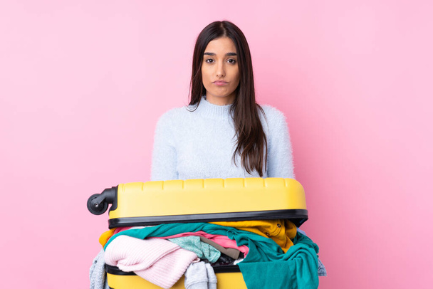 Traveler woman with a suitcase full of clothes over isolated pink background sad - Photo, Image