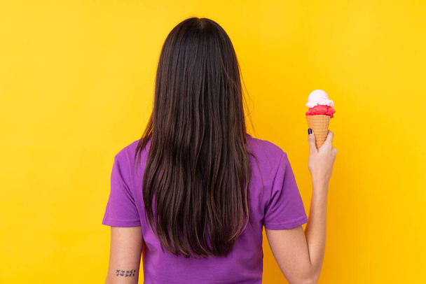 Young brunette woman with a cornet ice cream over isolated yellow background in back position - Fotografie, Obrázek