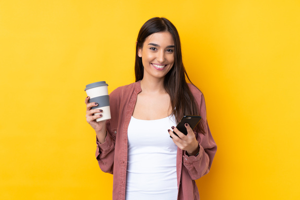 Young brunette woman over isolated yellow background holding coffee to take away and a mobile - Photo, Image