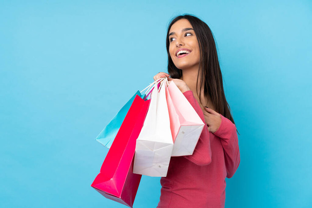 fiatal barna nő felett elszigetelt kék háttér gazdaság bevásárló táskák és mosolygós - Fotó, kép