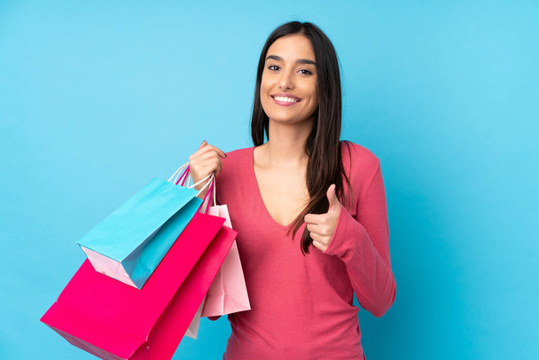 Young brunette woman over isolated blue background holding shopping bags and with thumb up - Фото, изображение
