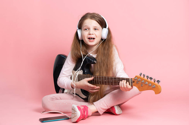 Music lessons: a schoolgirl learns to play the electric guitar. Portrait on a pink background. - Valokuva, kuva