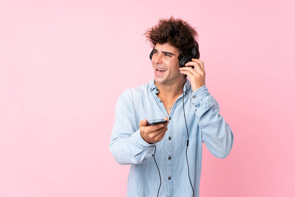 Joven hombre caucásico sobre fondo rosa aislado usando el móvil con auriculares y cantando
 - Foto, Imagen