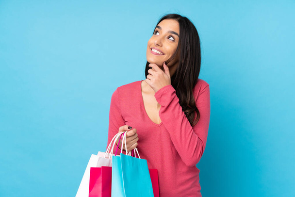 Mladá brunetka žena přes izolované modré pozadí držení nákupních tašek a myšlení - Fotografie, Obrázek