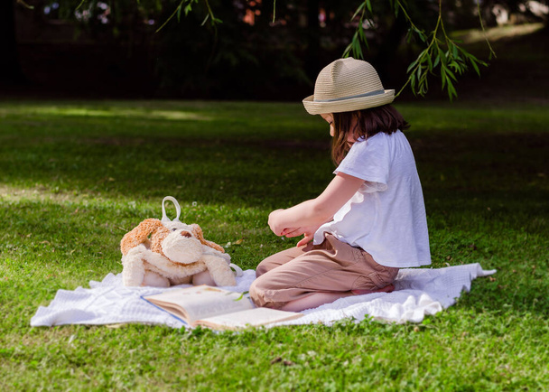 Ragazza prescolare con un cappello di paglia nel parco con libro. Bambino nel parco durante un picnic estivo
. - Foto, immagini