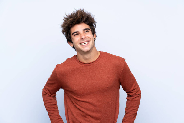 Jonge blanke man over geïsoleerde blauwe achtergrond poseren met armen op heup en glimlachen - Foto, afbeelding