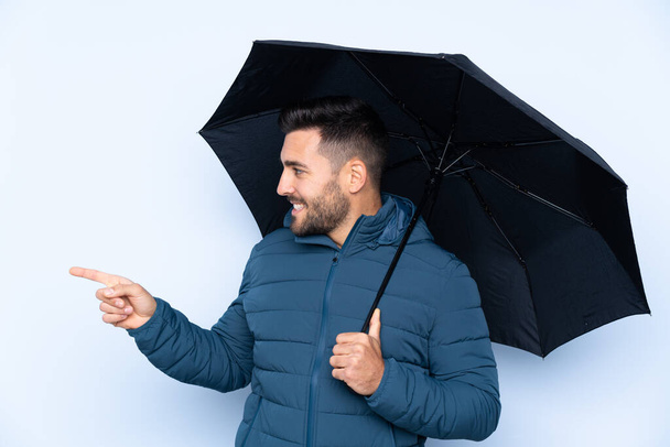 İzole edilmiş arka planda şemsiye tutan adam ürünü sunmak için kenarı işaret ediyor. - Fotoğraf, Görsel