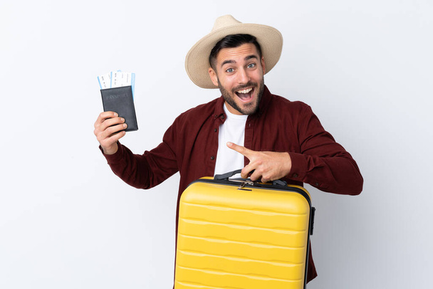Молодой красивый мужчина на изолированном белом фоне в отпуске с чемоданом и паспортом и удивлен
 - Фото, изображение