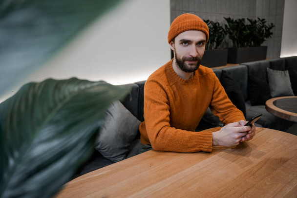 Hombre hipster llevado en naranja sentado en la cafetería y el sitio web de búsqueda en línea, la lectura en el teléfono móvil. Freelance. Comunicación en línea. Concepto tecnológico, usuario de Internet - Foto, Imagen