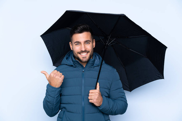 Άντρας κρατώντας μια ομπρέλα πάνω από απομονωμένο φόντο που δείχνει προς την πλευρά για να παρουσιάσει ένα προϊόν - Φωτογραφία, εικόνα