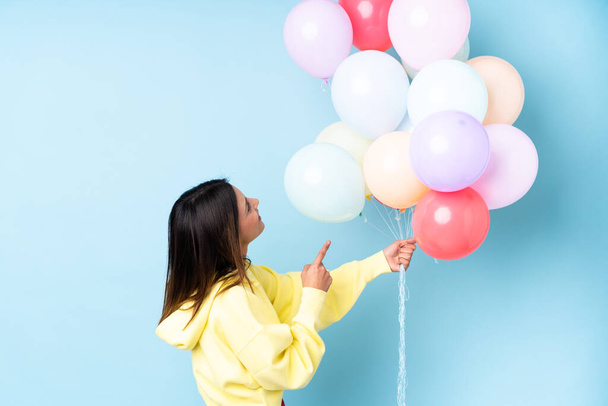 Woman holding balloons in a party over isolated blue background pointing back with the index finger - Photo, Image
