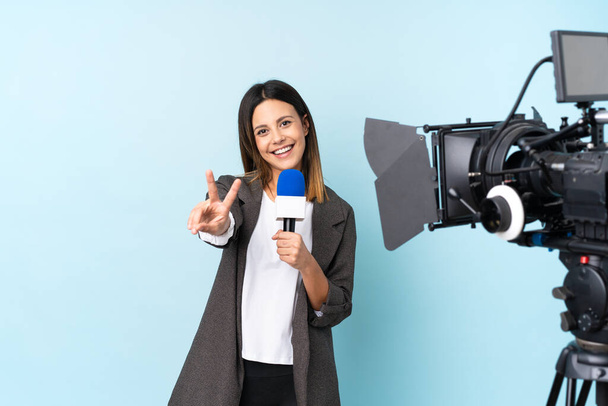 Женщина-репортер держит микрофон и сообщает новости на изолированном синем фоне улыбаясь и показывая знак победы
 - Фото, изображение